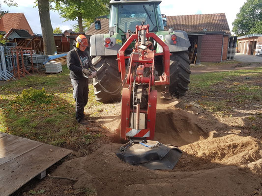 Wurzelfräse am Traktor arbeitet in einer großen Grube, Bediener steht in Schnittschutzkleidung daneben.